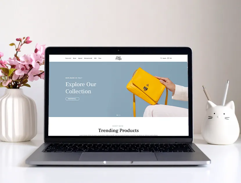 Mockup di un sito e-commerce visualizzato su laptop, su una scrivania minimal con decorazioni in ceramica bianca e fiori.